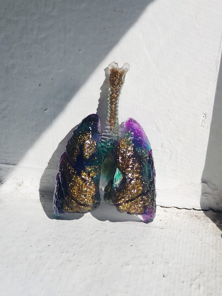 Gold Glitter Lungs Close up photo of an epoxy resin magnet sitting propped up in the sunlight. The magnet is gold glitter, purple, and teal, and shaped like human lungs.