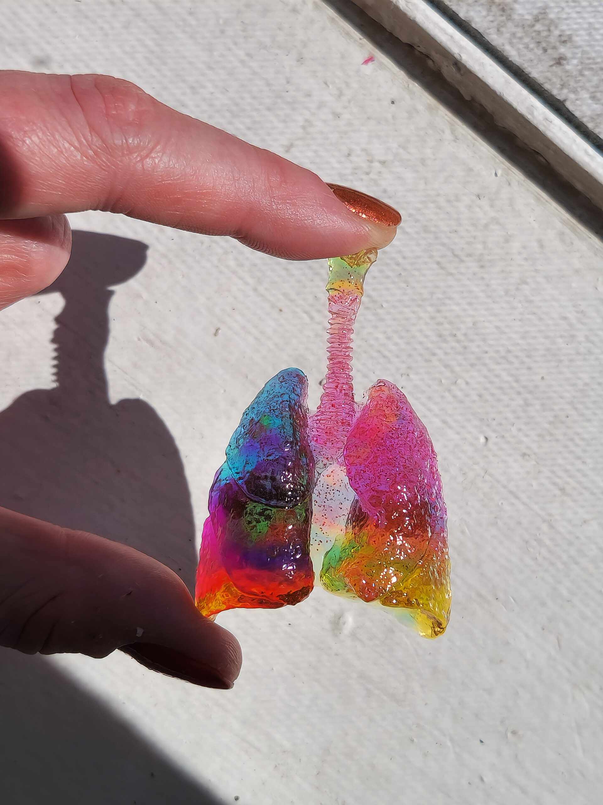 Glossy 3D human lungs held up in the sunlight, showing its translucent yellow, pink, and blue hue with holographic flecks.