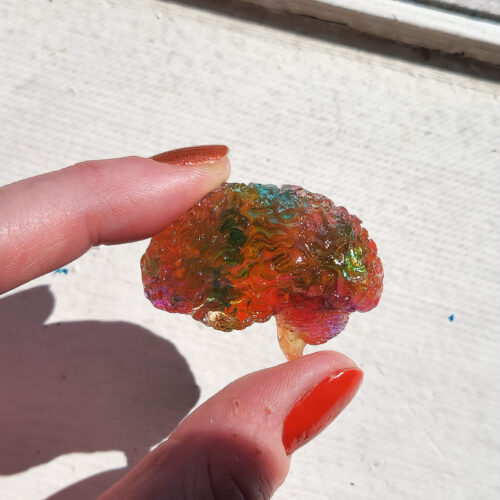 Glossy 3D human brain in side view, held up in the sunlight, showing its translucent yellowish pink hue with teal swatches and holographic flecks.