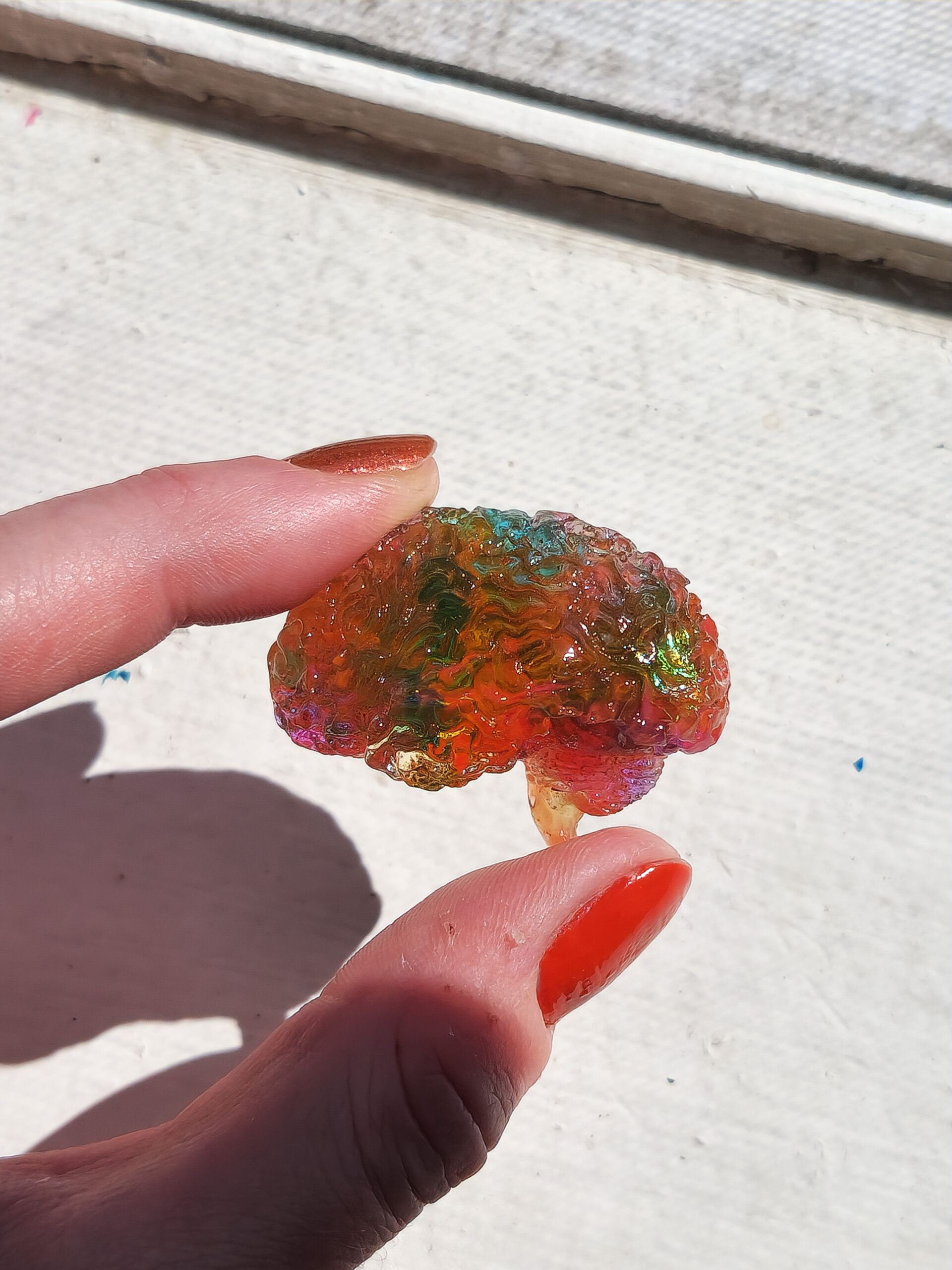 Glossy 3D human brain in side view, held up in the sunlight, showing its translucent yellowish pink hue with teal swatches and holographic flecks.