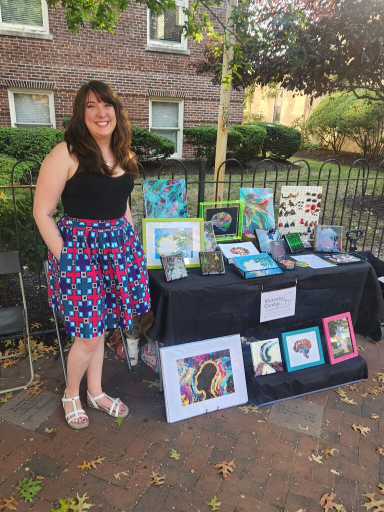 CollingswoodFirstFriday Victoria Corbit standing in front of a table covered with her art, hands in pockets of a pink and blue skirt, big smile on face.