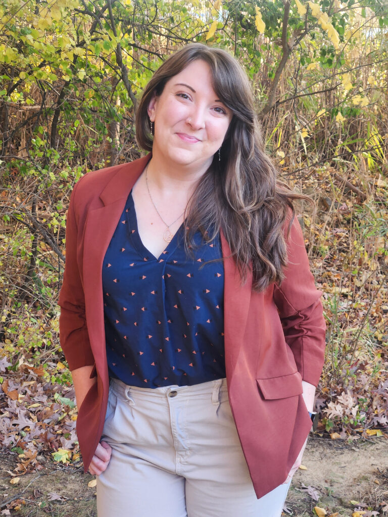 Headshot1 Victoria Corbit standing outside in a burnt orange blazer, dark teal blouse, and beige pants, hands in pockets and looking at the camera smiling.,