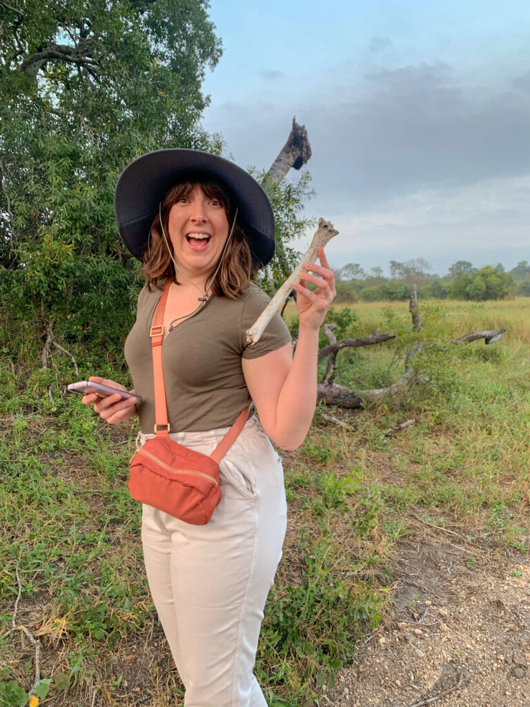 VCZimbabwe Victoria Corbit standing outside in an olive green v neck, beige pants, wide brim hat, and salmon colored cross body purse. She's holding up a femur bone with a huge open mouth grin on her face.