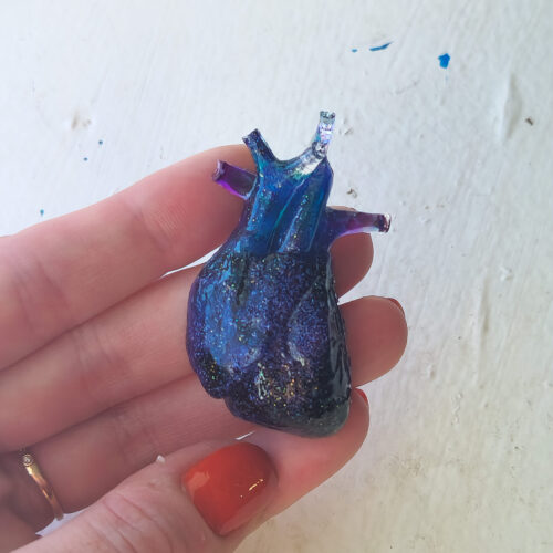3D anatomical heart with a glossy sheen held out in the sun. Heart is a deep blue and purple with glitter.